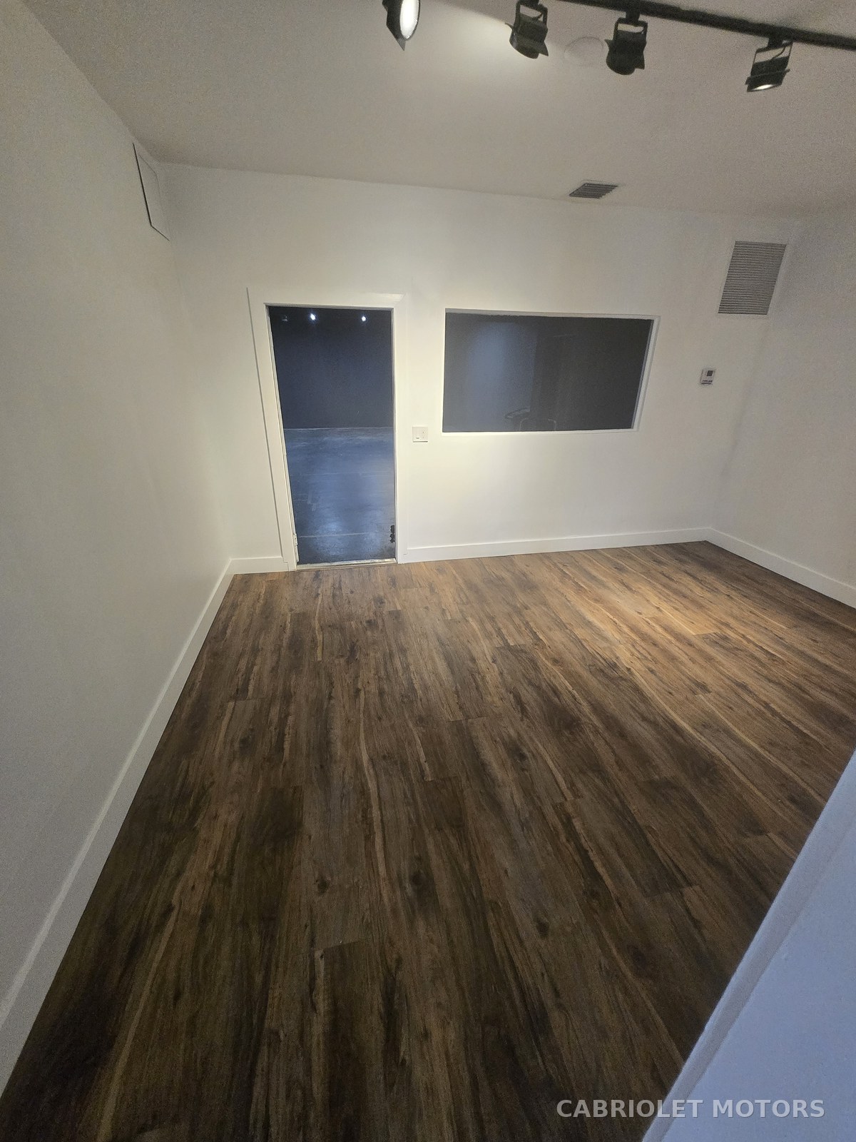 Owner's office before renovation, an empty white-walled room with wood-look plank flooring, track lighting and an interior observation window
